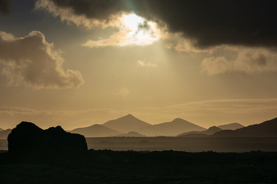 View of the volcanic landscape with mountain silhouettes under a dramatic sky with sunbeams breaking through clouds in Lanzarote, Canarias, Spain.