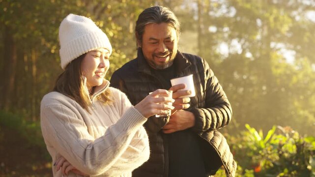 Asian couple drinking hot coffee at campsite on mountain peak and looking sea clouds during sunrise. Man and woman traveler enjoy camping outdoor lifestyle, relaxing in nature slow living in forest