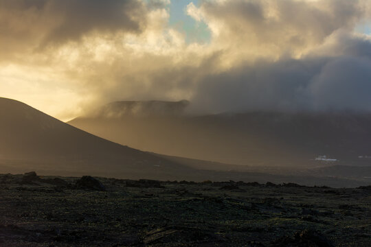 View of volcanic mountains partially covered by low clouds during golden hour over a rocky lava field in Lanzarote, Canarias, Spain.