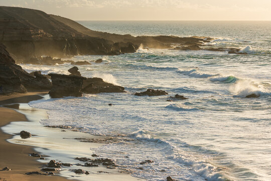 View of the rugged coastline of the Canary Islands with waves crashing against volcanic rocks and sandy beaches under a warm golden sunset light in Lanzarote, Canarias, Spain.
