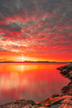 View of a vibrant sunset over the calm waters of a fjord with rocky shores and dramatic red clouds reflecting on the surface Nuuk, Sermersooq Municipali, Greenland.