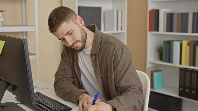 Man writing with blue pen at desktop computer and monitor, leaning forward with hand on keyboard, bookshelves nearby in building; focused productivity.