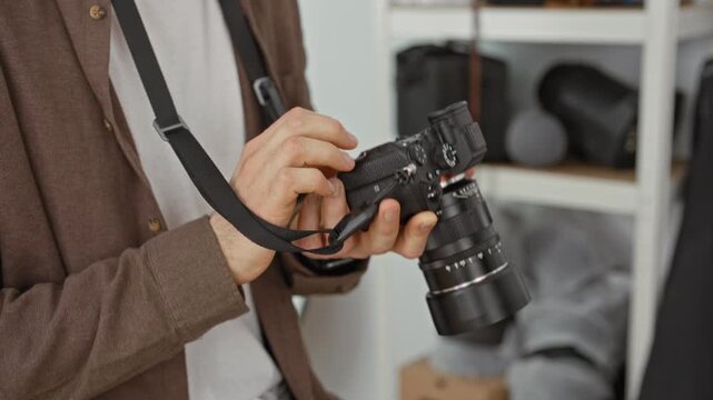 Young man adjusts camera dials while holding lens and strap in a photography studio with shelves of gear and softboxes visible; concentration craft.