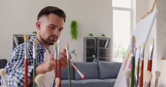 Artist and painter applies color with a brush to abstract painting on canvas set on easel in studio or at home workshop, with palette and paints. He adds details with bold strokes. Creative hobby.