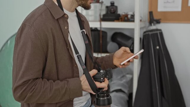 Young man holding smartphone and dslr camera, fingers tapping screen and grasping lens while wearing camera strap in studio; focus preparation.