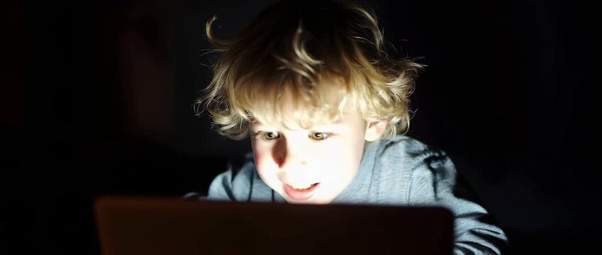 A little toddler in a gloomy setting is staring at a brilliant laptop screen. A child with wide eyes and an open mouth expressing astonishment and interest