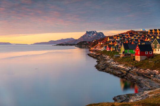 View of colorful houses along the rocky coastline with the Sermitsiaq mountain in the background during a vibrant sunset over the calm fjord in Nuuk, Sermersooq Municipality, Greenland.