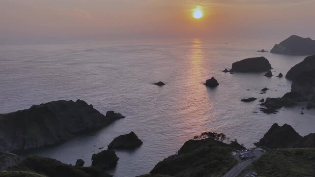 Aerial lateral drone shot of sunset over ocean with sun reflection path and coastal road at Aiai Misaki in Japan