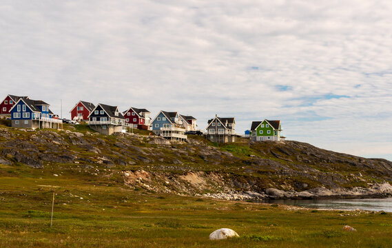 View of colorful wooden houses perched on a rocky ridge overlooking a grassy field and the sea under a cloudy sky in Nuuk, Sermersooq Municipality, Greenland.