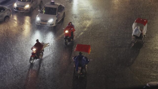 Two cyclos carrying passengers move through intense rain on busy Hanoi street with car headlights reflecting on soaked asphalt