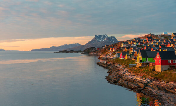 View of colorful wooden houses perched on a rocky shoreline overlooking the calm fjord waters under a soft sunset sky with distant mountains Nuuk, Sermersooq Municipali, Greenland.
