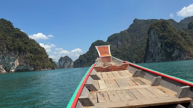 Wide view of a long-tail boat sailing towards massive limestone mountain ranges on Cheow Lan Lake. Stunning Avatar-like scenery and emerald waters in Khao Sok National Park, Southern Thailand.