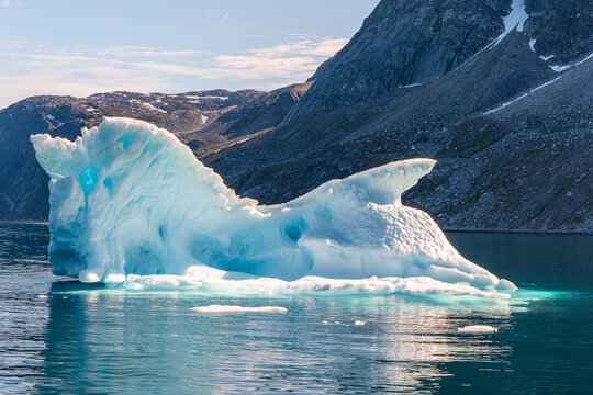 View of a massive blue iceberg with jagged peaks floating in the calm waters of a fjord surrounded by rocky mountains in Nuuk, Sermersooq Municipality, Greenland.