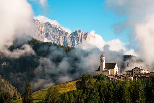 View of a small alpine village with a historic church nestled on a green hillside against the backdrop of the misty Dolomite mountains Trentino-South Tyrol, Italy.