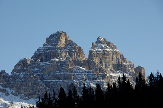 View of the snow-dusted jagged peaks of the Dolomites rising above a dark pine forest under a clear blue sky in Trentino-South Tyrol, Italy.