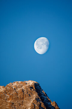 View of the gibbous moon in a clear blue sky above a rugged snow-dusted mountain peak in Trentino-South Tyrol, Italy.