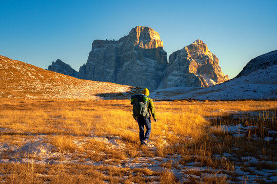 View of a hiker walking through golden alpine meadows towards the sunlit peaks of the Dolomites in Trentino-South Tyrol, Italy.