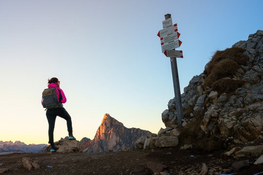 View of a female hiker with a backpack standing by a trail signpost looking at a sharp mountain peak during sunset in the Dolomites, Trentino-South Tyrol, Italy.
