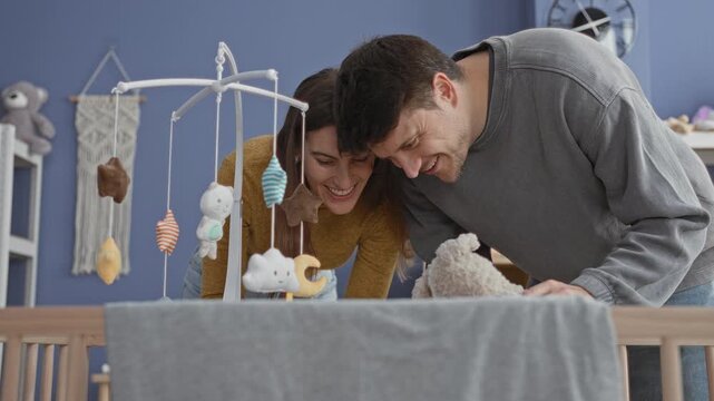 Young man and woman lean over crib, smiling and cradling baby under hanging mobile in nursery; family bond joy.