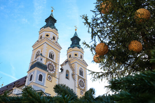 View of the twin bell towers of Brixen Cathedral with clocks and green spires behind a decorated Christmas tree under a blue sky in Trentino-South Tyrol, Italy.