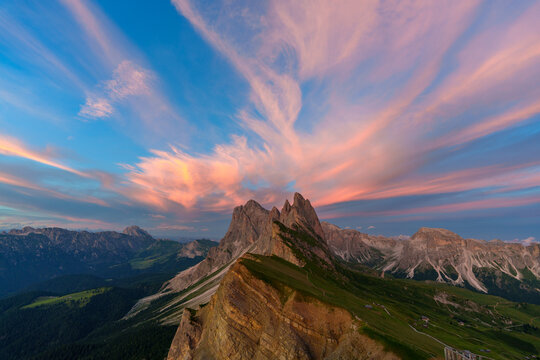 View of the jagged Seceda mountain peaks and green alpine meadows under a vibrant sunset sky with pink wispy clouds in Trentino-South Tyrol, Italy.