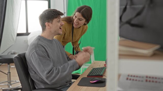 Young man cheering at keyboard while woman photographer leans over holding camera in studio with green screen and softbox lighting; collaboration.