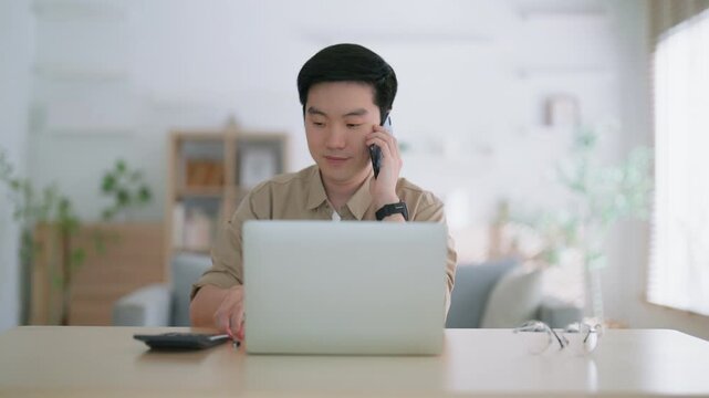 Happy young Asian man working on laptop in living room. Freelance worker male sitting at desk writing notes while watching webinar, studying online