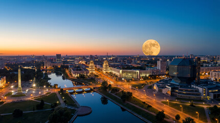 Obraz premium Panoramic view of a city skyline at dusk with a large full moon rising over the buildings and illuminated streets.