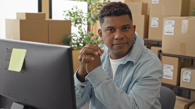 Man at computer clasping hands and smiling among stacked parcels and shipping boxes inside a packing building; friendly service.
