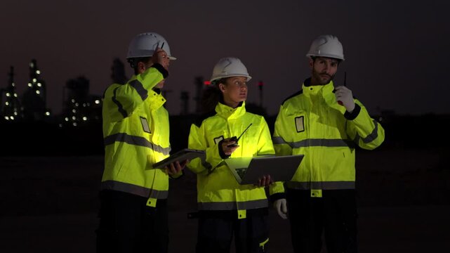 Workers in reflective jackets and helmets are talking to each other at night. They stand next to a vehicle, with the industrial area of ​​an oil refinery and its lights visible in the background