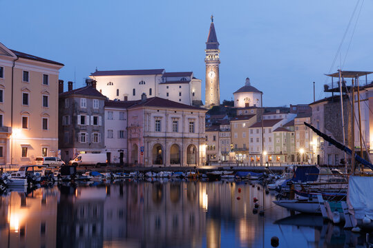 View of the harbor and St. George's Parish Church bell tower with illuminated historic buildings at dusk in Piran, Slovenia.