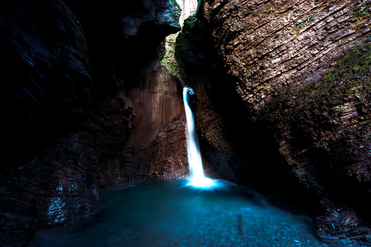 View of Kozjak Waterfall cascading into a turquoise pool within a dark limestone cavern in Slovenia.