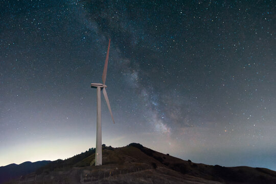 View of a wind turbine at Parco EOLICO Passo Cappelletta under a starry night sky with the Milky Way galaxy visible over the hills in Albareto, Emilia-Romagna, Italy.