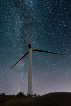 View of a tall wind turbine at Parco Eolico Passo Cappelletta under a bright starry night sky with the Milky Way galaxy Albareto, Emilia-Romagna, Italy.