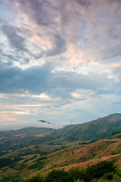 View of wind turbines of Parco Eolico Passo Cappelletta standing on rolling hills under a dramatic cloudy sky with morning mist in Albareto, Emilia-Romagna, Italy.