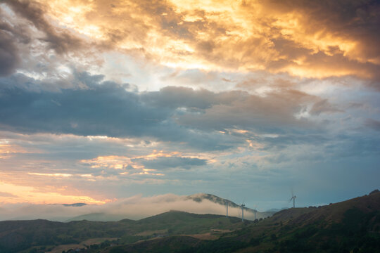 View of the wind turbines at Parco Eolico Passo Cappelletta on rolling green hills under a dramatic sunset sky with orange clouds and mist in Albareto, Emilia-Romagna, Italy.