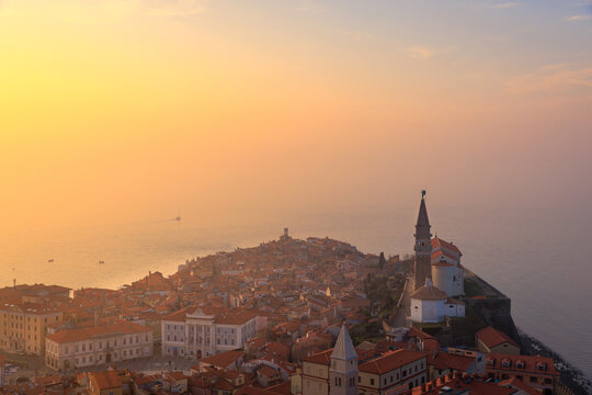 View of the historic old town of Piran with the St. George's Parish Church bell tower and red rooftops overlooking the Adriatic Sea during a hazy golden sunset in Slovenia.