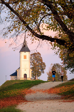 View of a white church on a hill with people jogging along a path covered in autumn leaves under a soft sunset sky in Slovenia.
