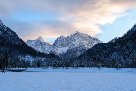 View of snow-covered alpine mountains and a frozen lake surrounded by pine trees under a soft winter sky Slovenia.