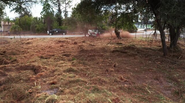 A piece of machinery stirs up dust while clearing a lot near a quiet road lined with trees. Useful for construction and land development visuals.