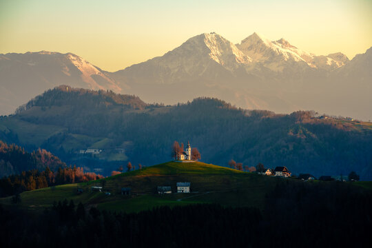 View of the historic Saint Thomas Church on a green hilltop with the snow-capped Julian Alps in the background during golden hour Slovenia.