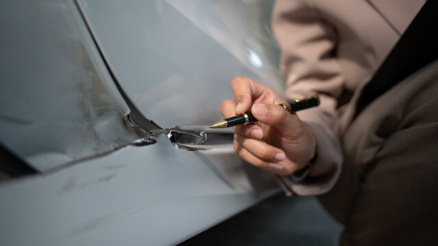 Close-up of a person using a pen to mark a damaged dent and scratch on a car body panel, preparing for auto body repair or insurance assessment.