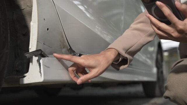 Close-up of a person inspecting a dent and scratch on a car bumper, holding a smartphone to document vehicle damage for insurance or repair estimate.