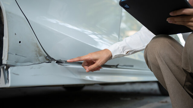 Close-up of a car&rsquo;s lower side panel with visible dent and paint damage as a person points to the scrape while holding a clipboard, suggesting vehicle inspection or insurance claim.