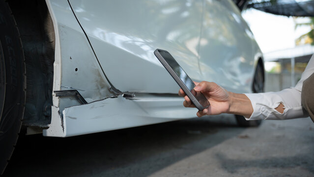 Close-up of damaged car rocker panel near wheel while a person photographs the dent with a smartphone for insurance claim or repair estimate outdoors.