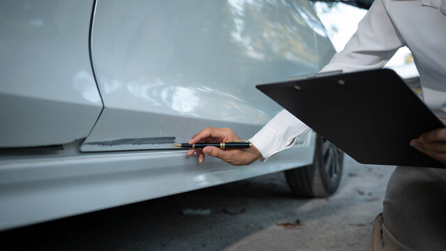 Close-up of car side with scratch damage as inspector writes notes on clipboard during vehicle assessment outdoors.