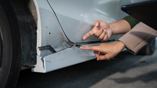 Close-up of hands pointing at scratched and dented car door and side panel near wheel, suggesting vehicle accident damage inspection and insurance claim repair concept.