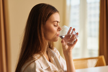Woman drinking water from a clear glass at home in soft morning light, relaxed profile portrait by a window, healthy hydration routine, calm atmosphere and casual lifestyle moment. © SHOTPRIME STUDIO