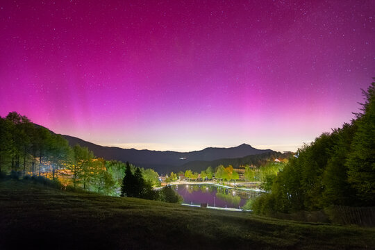 View of Passo del Cerreto under a vibrant pink aurora borealis with a starry night sky, mountain silhouettes, and a small lake reflecting the colorful sky Fivizzano, Tuscany, Italy.