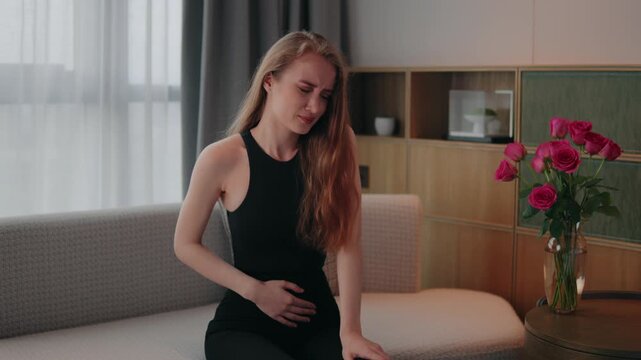 Young woman suffering menstruation, sits on a cozy sofa at home, grimacing and holding her lower back, abdomen in pain from severe backache and stomach cramps during illness, menstruation